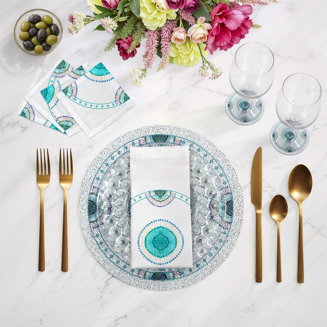 Dining table setting with decorative plates, cutlery, and flowers on a marble surface.